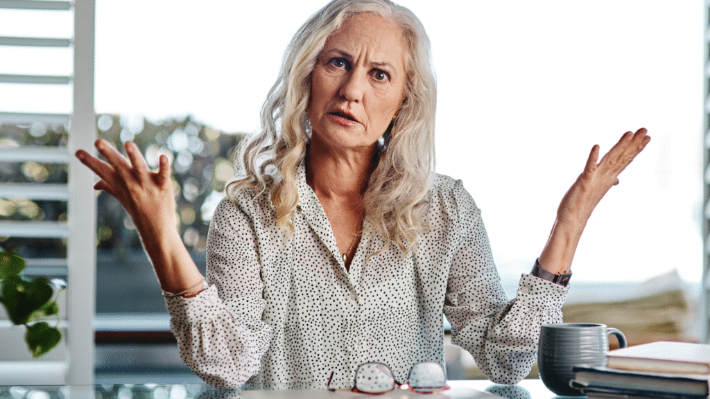 Woman shrugging with a confused expression, sitting at a desk with reading glasses and a mug in front of her