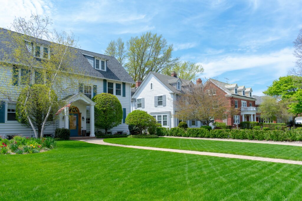 Charming residential street with traditional homes, manicured lawns, and bright spring greenery under a blue sky.
