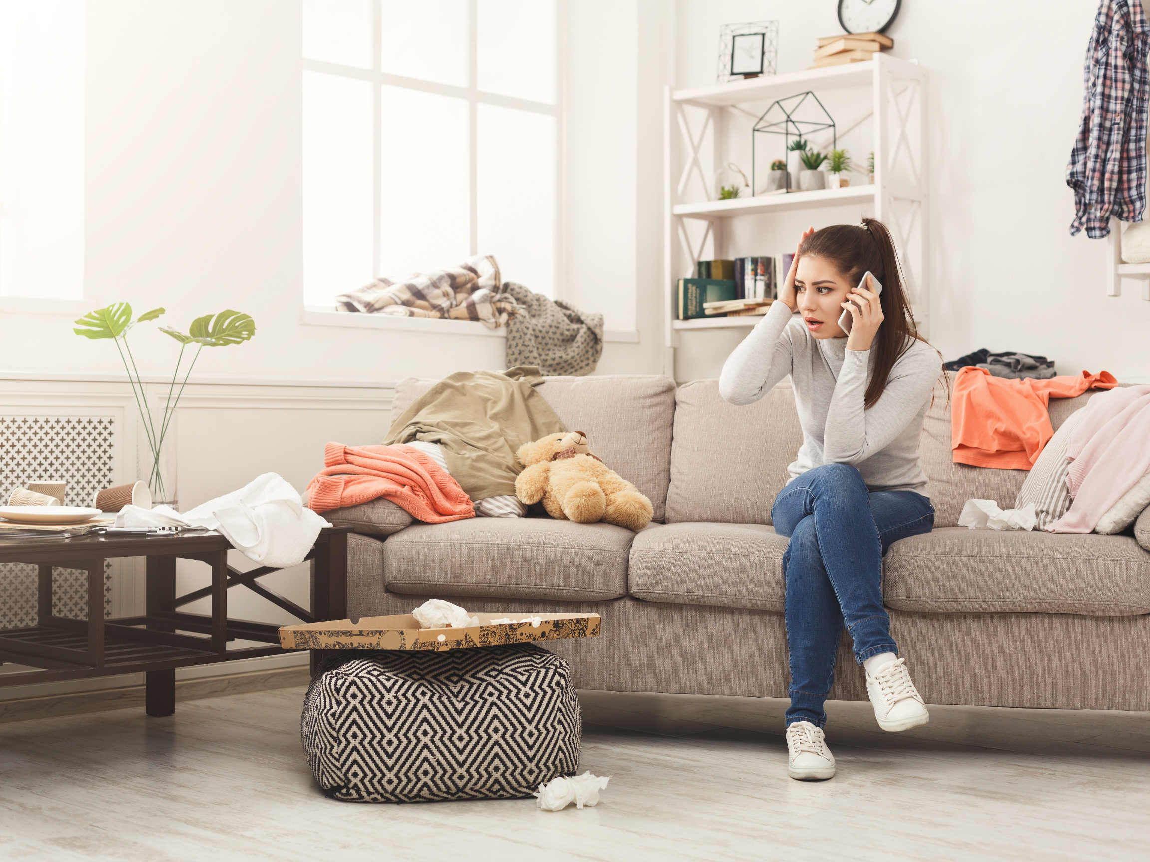 Woman on the phone in an untidy living room, looking worried about her messy house.