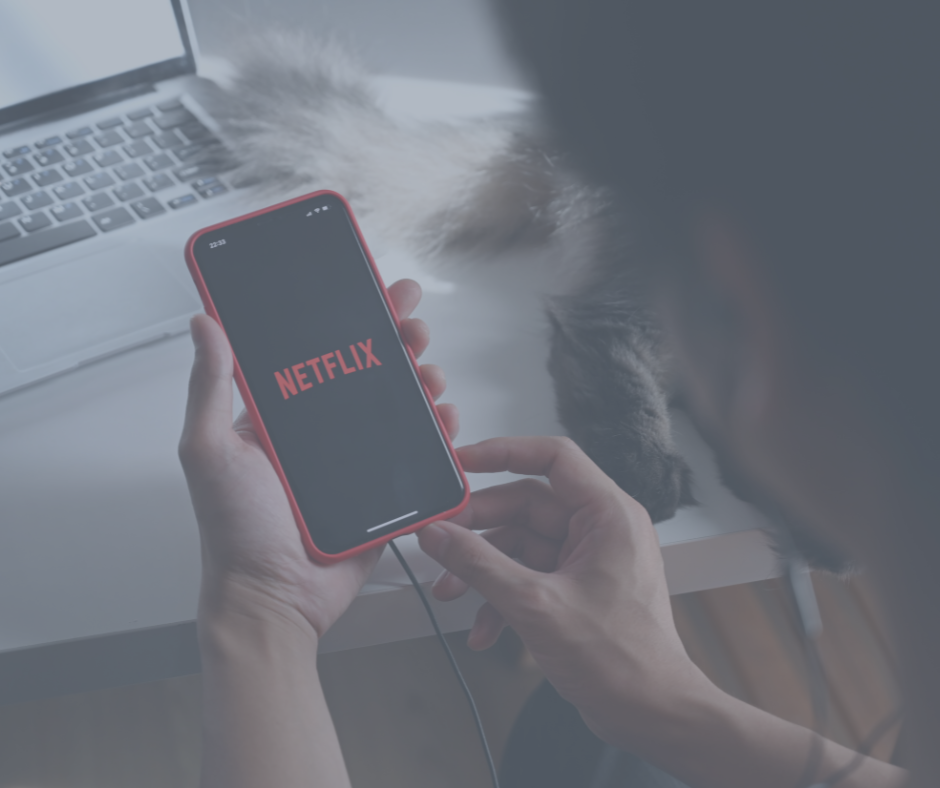 Person holding a smartphone with Netflix app open, sitting at a desk with a laptop nearby