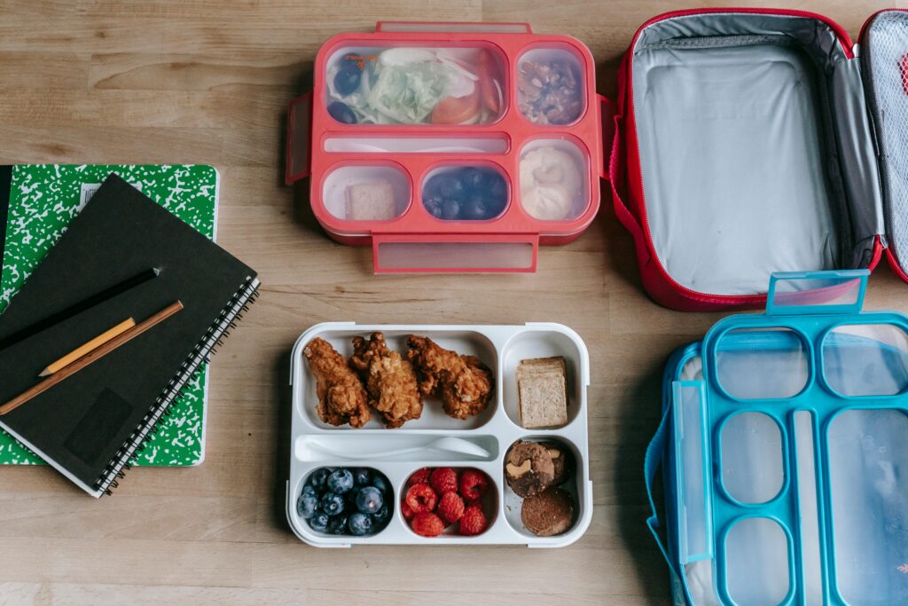 Overhead view of packed school lunch containers, fruit, fried chicken, and notebooks, symbolizing the back-to-school routine I’m happy to leave behind.