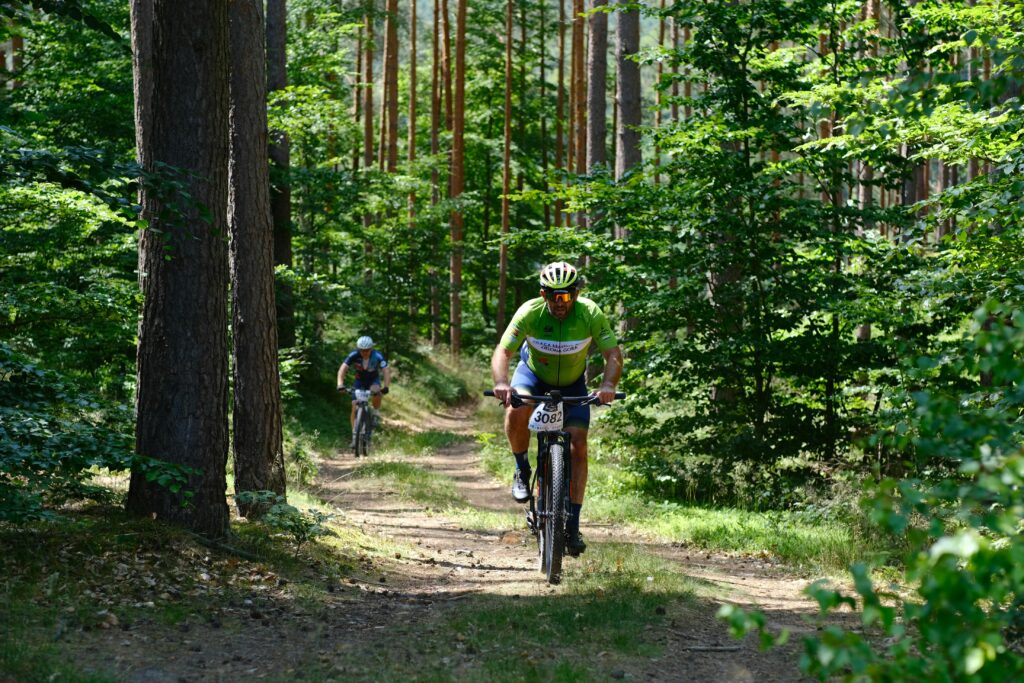Two cyclists riding along a sunlit forest trail, one in a bright green jersey leading, symbolizing the shift from relaxed mornings to active, ambitious plans.