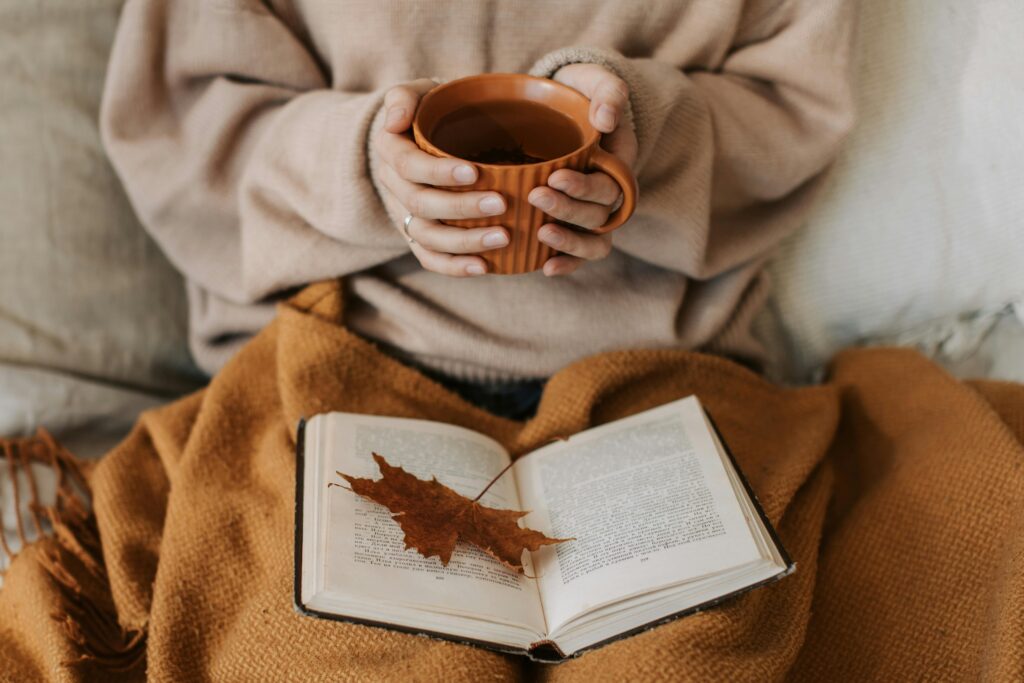 Close-up of a person wrapped in a warm brown blanket, holding a terracotta mug of tea, with an open book and a dried maple leaf resting on the pages.