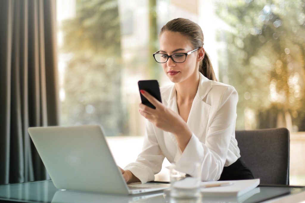 Woman in glasses checking her phone while working on a laptop, representing clear communication during the home selling process.