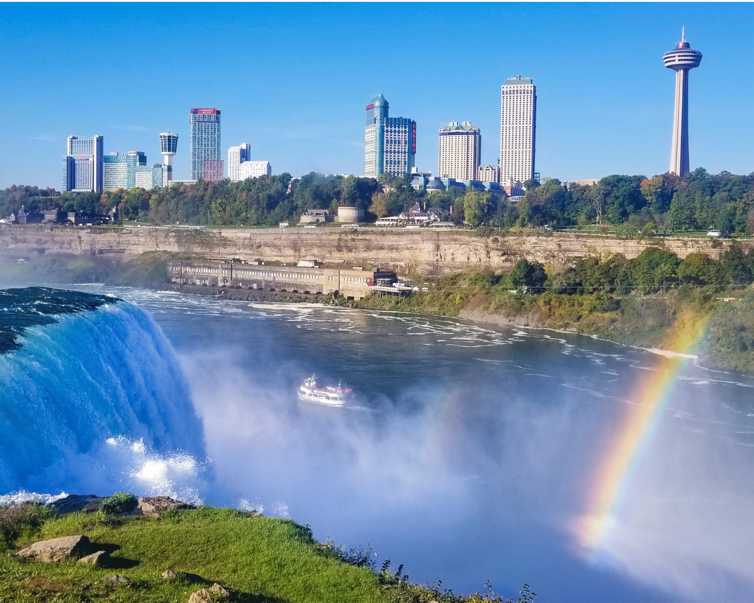 View of Niagara Falls with the city skyline and a rainbow over the water.